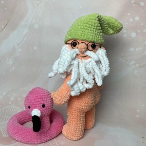 May include: A crocheted gnome doll with a green hat, white beard, and orange body. The gnome is holding a pink flamingo pool float.