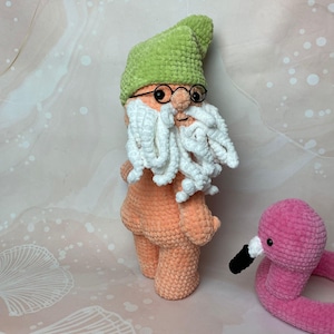 May include: A crocheted gnome with a green hat, white beard, and glasses stands next to a pink crocheted flamingo.