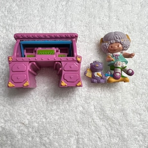 May include: A vintage toy set featuring a purple desk with drawers and a small figurine of a girl with a bear. The desk has a blue interior and gold accents. The girl is wearing a green top and has a bow.