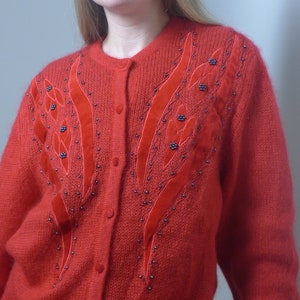 Puede incluir: Un c&aacute;rdigan de punto rojo con un dise&ntilde;o floral con cuentas negras y detalles de terciopelo rojo.