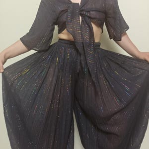 May include: A two-piece black outfit with a tie-front top and wide-leg pants. The fabric has a subtle shimmer with iridescent threads. The top has a V-neck and flared sleeves. The pants have a drawstring waist and a flowing silhouette.