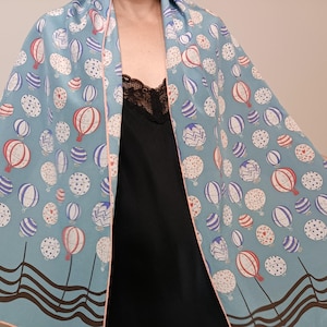 May include: A light blue scarf with a repeating pattern of hot air balloons in red, white, and blue. The scarf has a pink border and a brown wavy design at the bottom. The scarf is draped over a black top.