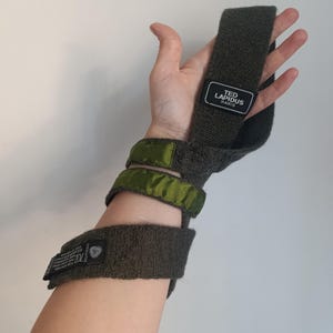 May include: A dark gray knit wrist accessory featuring two olive green satin bands and a black rectangular label with the white text TED LAPIDUS PARIS. The accessory includes a secondary black label with white text detailing fabric composition.