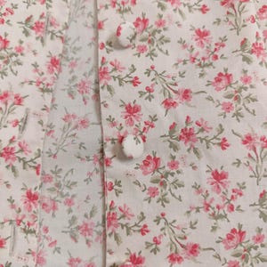K&ouml;nnte beinhalten: Nahaufnahme eines wei&szlig;en Stoffes mit einem zarten Blumenmuster in Rosa und Gr&uuml;n. Der Stoff zeigt kleine, detaillierte Bl&uuml;ten und gr&uuml;ne Bl&auml;tter. Drei wei&szlig;e, stoffbezogene Kn&ouml;pfe sind sichtbar, was auf ein Kleidungsst&uuml;ck oder Textilartikel hindeutet.
