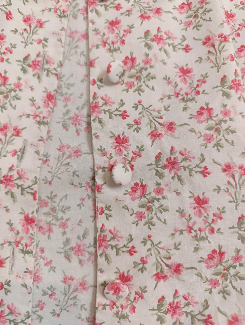 K&ouml;nnte beinhalten: Nahaufnahme eines wei&szlig;en Stoffes mit einem zarten Blumenmuster in Rosa und Gr&uuml;n. Der Stoff zeigt kleine, detaillierte Bl&uuml;ten und gr&uuml;ne Bl&auml;tter. Drei wei&szlig;e, stoffbezogene Kn&ouml;pfe sind sichtbar, was auf ein Kleidungsst&uuml;ck oder Textilartikel hindeutet.