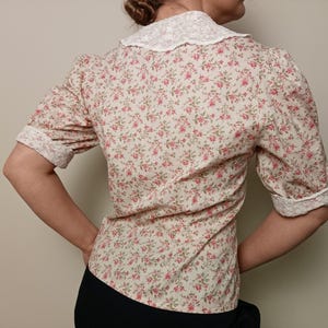 K&ouml;nnte beinhalten: Eine Bluse mit floralem Muster und einem wei&szlig;en Spitzenkragen und -b&uuml;ndchen. Das kurz&auml;rmelige Hemd hat ein Muster aus kleinen rosa Blumen auf cremefarbenem Hintergrund. Die Manschetten und der Kragen sind mit wei&szlig;er Spitze verziert.