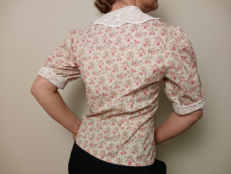 K&ouml;nnte beinhalten: Eine Bluse mit floralem Muster und einem wei&szlig;en Spitzenkragen und -b&uuml;ndchen. Das kurz&auml;rmelige Hemd hat ein Muster aus kleinen rosa Blumen auf cremefarbenem Hintergrund. Die Manschetten und der Kragen sind mit wei&szlig;er Spitze verziert.