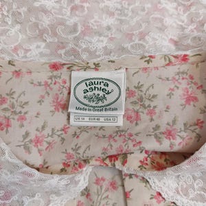 K&ouml;nnte beinhalten: Bluse mit floralem Muster und wei&szlig;em Spitzenkragen. Der Stoff zeigt ein Muster aus kleinen rosa Bl&uuml;ten und gr&uuml;nen Bl&auml;ttern auf cremefarbenem Hintergrund. Das Etikett zeigt "Laura Ashley" und die Gr&ouml;&szlig;en UK 14, EUR 40 und USA 12.