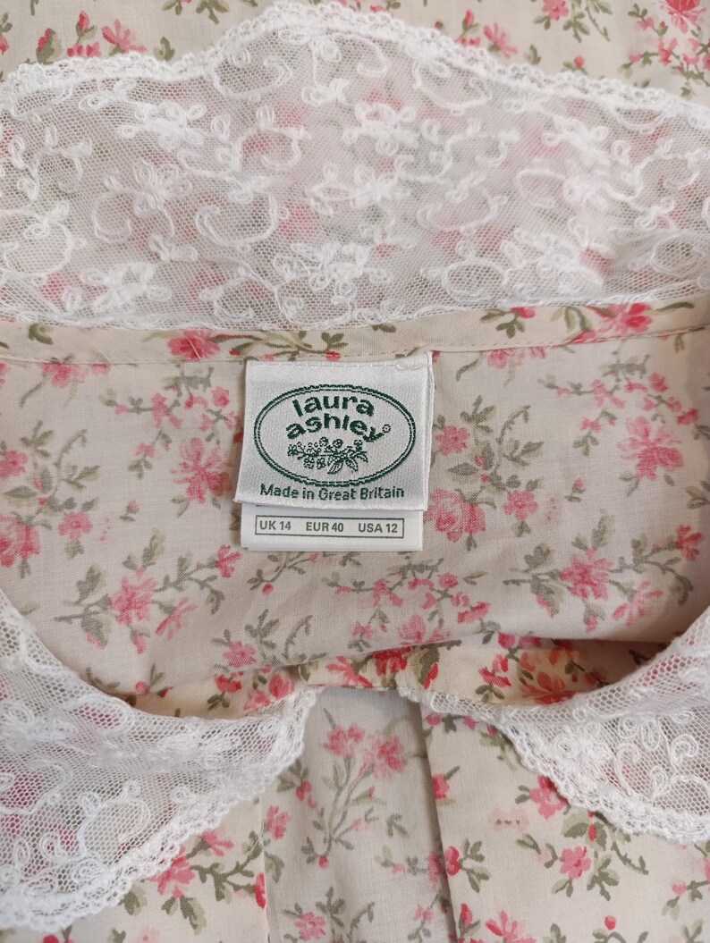 K&ouml;nnte beinhalten: Bluse mit floralem Muster und wei&szlig;em Spitzenkragen. Der Stoff zeigt ein Muster aus kleinen rosa Bl&uuml;ten und gr&uuml;nen Bl&auml;ttern auf cremefarbenem Hintergrund. Das Etikett zeigt "Laura Ashley" und die Gr&ouml;&szlig;en UK 14, EUR 40 und USA 12.