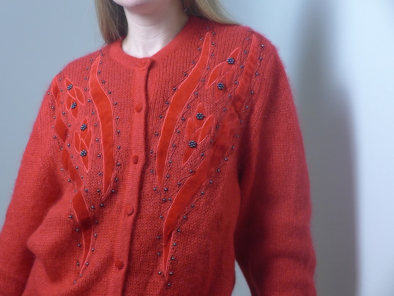 Puede incluir: Un c&aacute;rdigan de punto rojo con un dise&ntilde;o floral con cuentas negras y detalles de terciopelo rojo.
