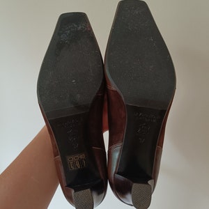 May include: A pair of brown suede heeled boots with a square toe. The boots have a black rubber sole and a small gold label on the back of each boot.