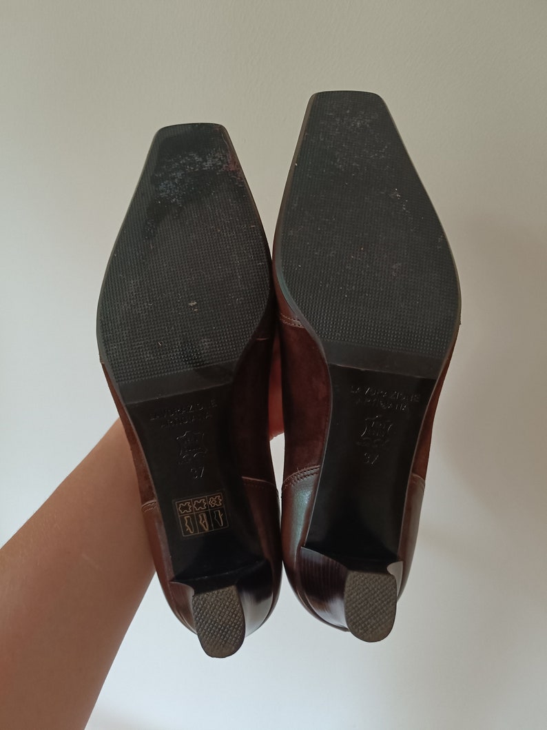 May include: A pair of brown suede heeled boots with a square toe. The boots have a black rubber sole and a small gold label on the back of each boot.