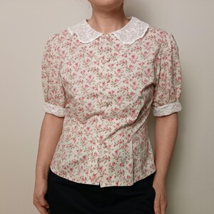 K&ouml;nnte beinhalten: Eine Bluse mit floralem Muster, wei&szlig;em Spitzenkragen und hochgekrempelten &Auml;rmeln. Das kurz&auml;rmelige Hemd hat ein Muster aus kleinen rosa Blumen auf cremefarbenem Hintergrund. Die Bluse ist vorne gekn&ouml;pft und hat eine taillierte Silhouette.
