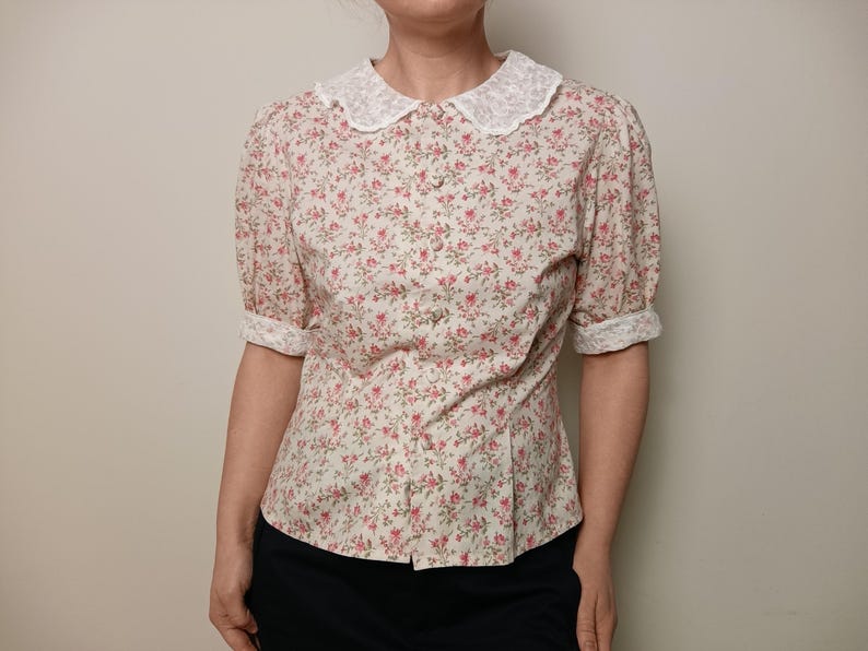 K&ouml;nnte beinhalten: Eine Bluse mit floralem Muster, wei&szlig;em Spitzenkragen und hochgekrempelten &Auml;rmeln. Das kurz&auml;rmelige Hemd hat ein Muster aus kleinen rosa Blumen auf cremefarbenem Hintergrund. Die Bluse ist vorne gekn&ouml;pft und hat eine taillierte Silhouette.