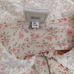 K&ouml;nnte beinhalten: Nahaufnahme eines Stoffes mit Blumenmuster und wei&szlig;er Spitzenborte. Der Stoff hat einen cremefarbenen Hintergrund mit kleinen rosa Bl&uuml;ten und gr&uuml;nen Bl&auml;ttern. Ein Etikett lautet "100% Cotton" und "BL 756".