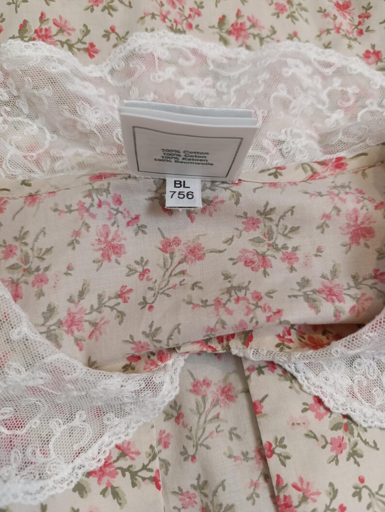 K&ouml;nnte beinhalten: Nahaufnahme eines Stoffes mit Blumenmuster und wei&szlig;er Spitzenborte. Der Stoff hat einen cremefarbenen Hintergrund mit kleinen rosa Bl&uuml;ten und gr&uuml;nen Bl&auml;ttern. Ein Etikett lautet "100% Cotton" und "BL 756".