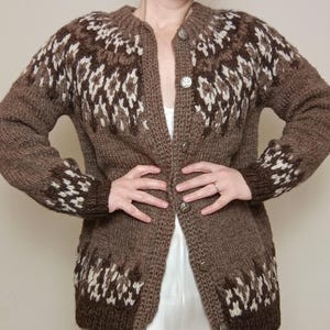 May include: A brown, button-up cardigan sweater with a geometric pattern in shades of brown and white. The long-sleeved sweater has a round neckline and is made of a thick, textured knit fabric. The sweater is approximately 76 cm long.