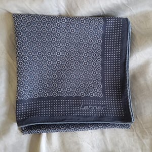 May include: A folded navy blue pocket square with a repeating floral pattern in light blue and white. The border features a dotted design. The brand name "Lehner" is printed in white.