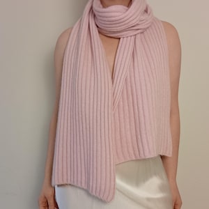 May include: A light pink ribbed scarf is draped around the neck and shoulders. The scarf is made of a soft material and has a vertical ribbed texture. The scarf is a solid color and is worn over a white satin top.