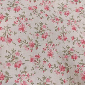 K&ouml;nnte beinhalten: Nahaufnahme eines Stoffes mit floralem Muster. Der Stoff ist wei&szlig; mit einem sich wiederholenden Muster aus rosa Rosen und gr&uuml;nen Bl&auml;ttern. Am oberen Bildrand ist eine wei&szlig;e Spitzentrimmung zu sehen.