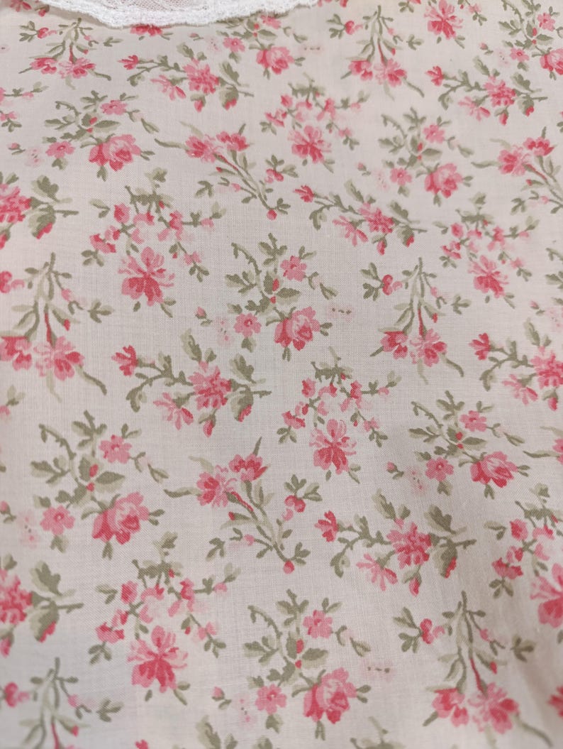 K&ouml;nnte beinhalten: Nahaufnahme eines Stoffes mit floralem Muster. Der Stoff ist wei&szlig; mit einem sich wiederholenden Muster aus rosa Rosen und gr&uuml;nen Bl&auml;ttern. Am oberen Bildrand ist eine wei&szlig;e Spitzentrimmung zu sehen.