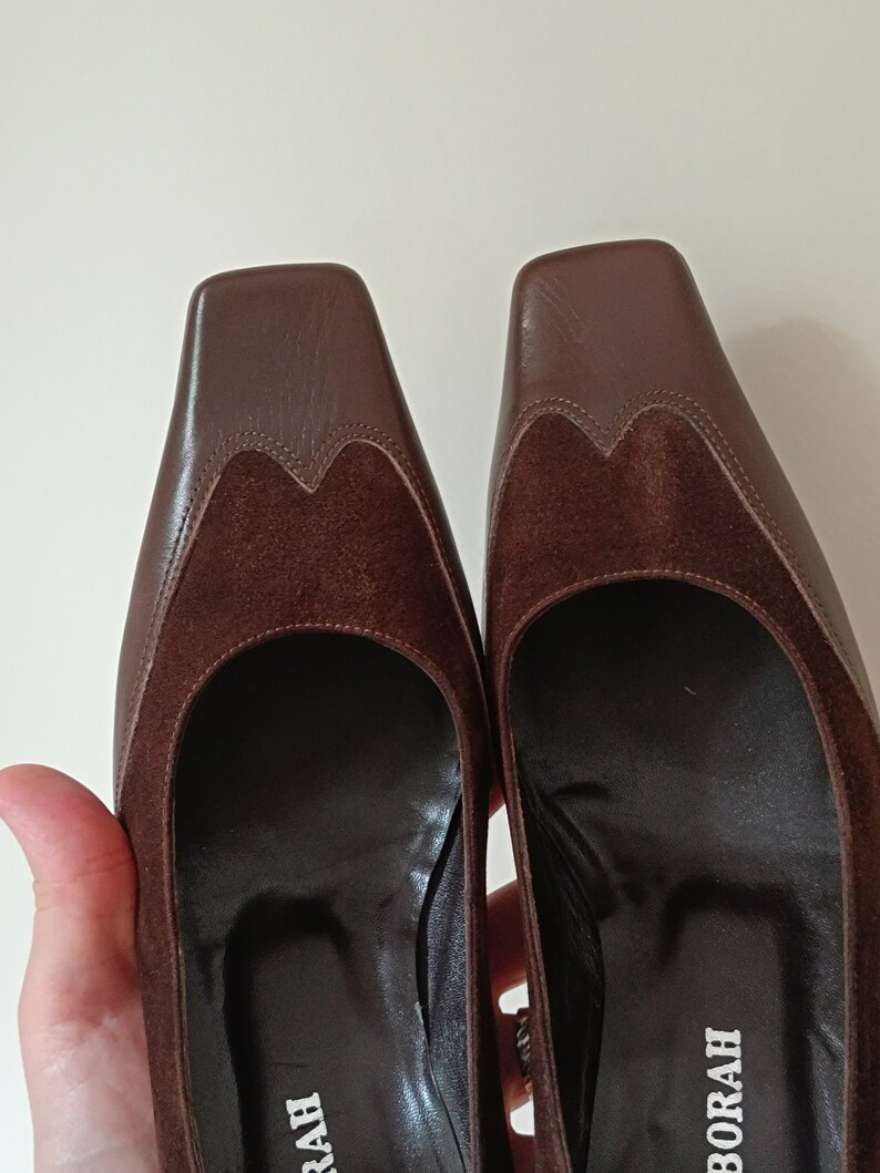 May include: A pair of brown leather heeled shoes with a suede detail on the toe. The shoes have a square toe and a low heel. The brand name "BORAH" is visible on the sole of one shoe.