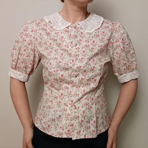 K&ouml;nnte beinhalten: Cremefarbene Bluse mit floralem Muster, wei&szlig;em Spitzenkragen und kurzen &Auml;rmeln. Die Bluse hat eine Knopfleiste vorne und eine taillierte Silhouette. Der Stoff ist mit kleinen rosa Blumen bedruckt.