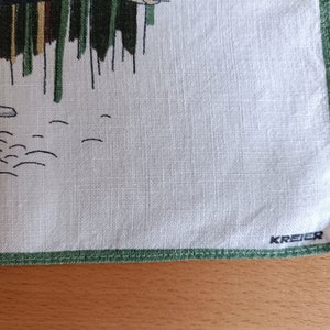 May include: A white linen tea towel with a green border and a green and brown illustration of a bird and reeds. The towel has the word "KREYER" printed in black in the bottom right corner.