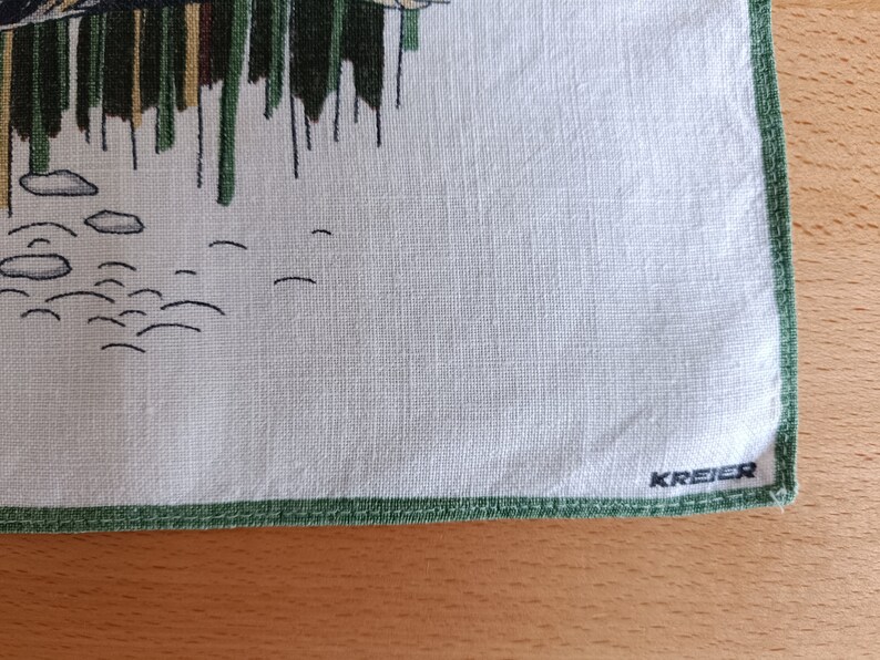 May include: A white linen tea towel with a green border and a green and brown illustration of a bird and reeds. The towel has the word "KREYER" printed in black in the bottom right corner.