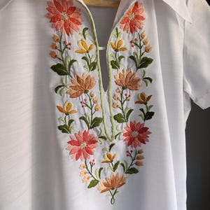 Op de afbeelding: Een wit shirt met korte mouwen, een puntige kraag en een diepe V-hals. Het kledingstuk is versierd met gedetailleerd bloemenborduursel in koraal, perzik, geel en groen, dat vanaf de halslijn naar beneden over de voorzijde is aangebracht.