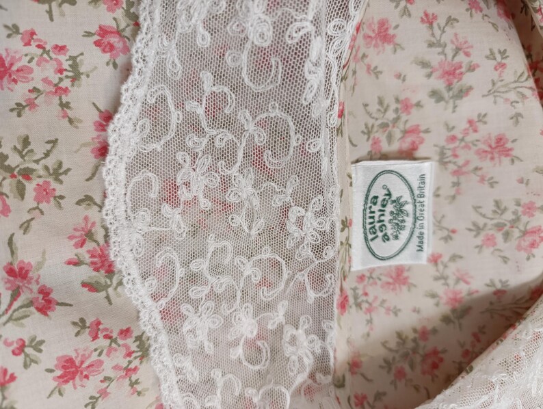 K&ouml;nnte beinhalten: Nahaufnahme eines floralen Stoffes mit wei&szlig;er Spitzenborte. Der Stoff hat ein Muster aus kleinen rosa Blumen und gr&uuml;nen Bl&auml;ttern auf cremefarbenem Hintergrund. Ein Etikett lautet "laura ashley Made in Great Britain."