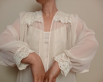 May include: A sheer, ivory-colored robe with delicate lace trim. The robe features long, flowing sleeves and lace cuffs. The neckline and shoulders are also adorned with lace. The garment appears to be made of a lightweight, possibly silk or chiffon material.