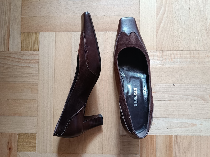 May include: A pair of brown leather heeled shoes with a suede detail on the toe. The shoes have a square toe and a block heel. The brand name "DEBORAH" is printed on the insole of one shoe.