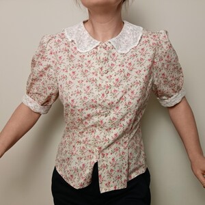K&ouml;nnte beinhalten: Bluse mit floralem Muster, wei&szlig;em Spitzenkragen und kurzen Puff&auml;rmeln. Die Bluse hat ein Muster aus kleinen rosa Blumen auf cremefarbenem Hintergrund und ist vorne gekn&ouml;pft.