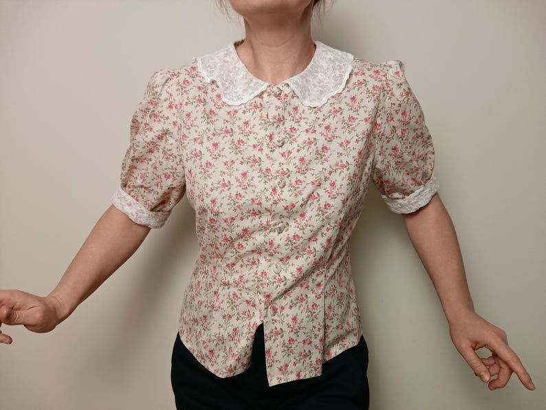 K&ouml;nnte beinhalten: Bluse mit floralem Muster, wei&szlig;em Spitzenkragen und kurzen Puff&auml;rmeln. Die Bluse hat ein Muster aus kleinen rosa Blumen auf cremefarbenem Hintergrund und ist vorne gekn&ouml;pft.