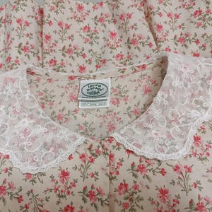 K&ouml;nnte beinhalten: Ein Kleidungsst&uuml;ck mit floralem Muster und wei&szlig;em Spitzenkragen. Der Stoff zeigt ein Muster aus kleinen rosa Bl&uuml;ten auf cremefarbenem Hintergrund. Der Kragen ist gewellt und verleiht dem Design eine zarte Note. Das Etikett lautet "Laura Ashley".