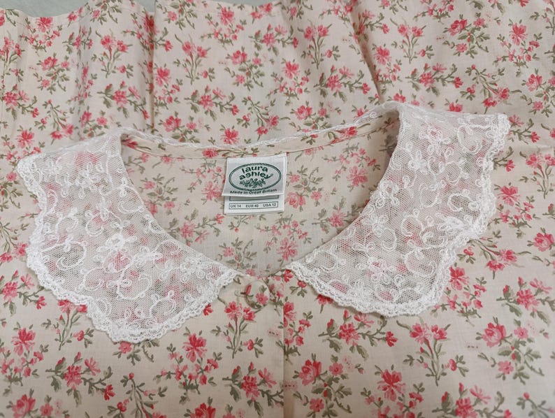 K&ouml;nnte beinhalten: Ein Kleidungsst&uuml;ck mit floralem Muster und wei&szlig;em Spitzenkragen. Der Stoff zeigt ein Muster aus kleinen rosa Bl&uuml;ten auf cremefarbenem Hintergrund. Der Kragen ist gewellt und verleiht dem Design eine zarte Note. Das Etikett lautet "Laura Ashley".