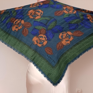 Könnte beinhalten: Ein quadratisches, teal-farbenes Tuch mit einem Blumenmuster aus orangefarbenen Rosen und blauen Blättern. Das Tuch hat einen grünen Rand mit strukturiertem Design und Fransen. Der Markenname "LANVIN" ist sichtbar.