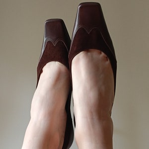 May include: A pair of brown suede and leather heeled shoes with a square toe. The shoes are worn and the toes are pointed upwards.