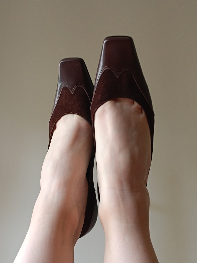 May include: A pair of brown suede and leather heeled shoes with a square toe. The shoes are worn and the toes are pointed upwards.