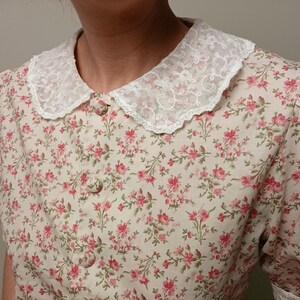 K&ouml;nnte beinhalten: Eine cremefarbene Bluse mit floralem Muster und wei&szlig;em Spitzenkragen. Das kurz&auml;rmelige Oberteil hat ein Muster aus kleinen rosa Blumen und gr&uuml;nen Bl&auml;ttern. Die Bluse hat eine Knopfleiste vorne und ein zartes, feminines Design.