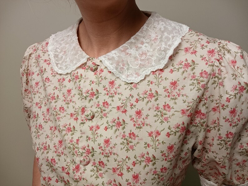 K&ouml;nnte beinhalten: Eine cremefarbene Bluse mit floralem Muster und wei&szlig;em Spitzenkragen. Das kurz&auml;rmelige Oberteil hat ein Muster aus kleinen rosa Blumen und gr&uuml;nen Bl&auml;ttern. Die Bluse hat eine Knopfleiste vorne und ein zartes, feminines Design.