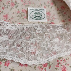 K&ouml;nnte beinhalten: Nahaufnahme eines floralen Stoffes mit wei&szlig;er Spitzenborte. Der Stoff hat ein Muster aus kleinen rosa Blumen auf cremefarbenem Hintergrund. Ein Etikett zeigt "Laura Ashley" und "Made in Great Britain".