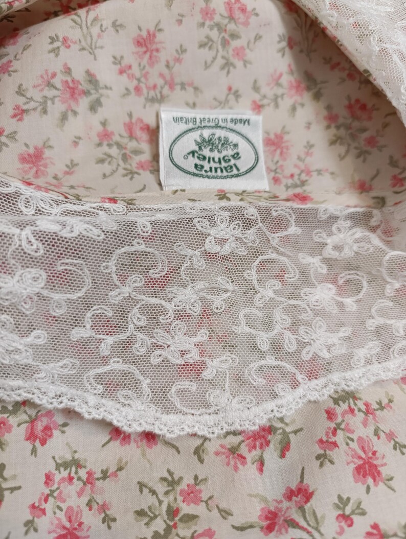 K&ouml;nnte beinhalten: Nahaufnahme eines floralen Stoffes mit wei&szlig;er Spitzenborte. Der Stoff hat ein Muster aus kleinen rosa Blumen auf cremefarbenem Hintergrund. Ein Etikett zeigt "Laura Ashley" und "Made in Great Britain".