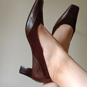 May include: A pair of brown leather heeled shoes with a square toe. The shoes have a suede panel on the back of the heel.