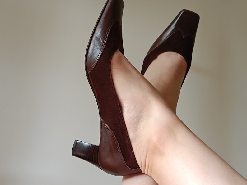 May include: A pair of brown leather heeled shoes with a square toe. The shoes have a suede panel on the back of the heel.
