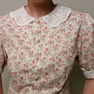 K&ouml;nnte beinhalten: Eine Bluse mit floralem Muster und einem wei&szlig;en Spitzenkragen und -b&uuml;ndchen. Die Bluse hat ein Muster aus kleinen rosa Blumen auf cremefarbenem Hintergrund. Sie hat kurze, gepuffte &Auml;rmel und eine Knopfleiste.