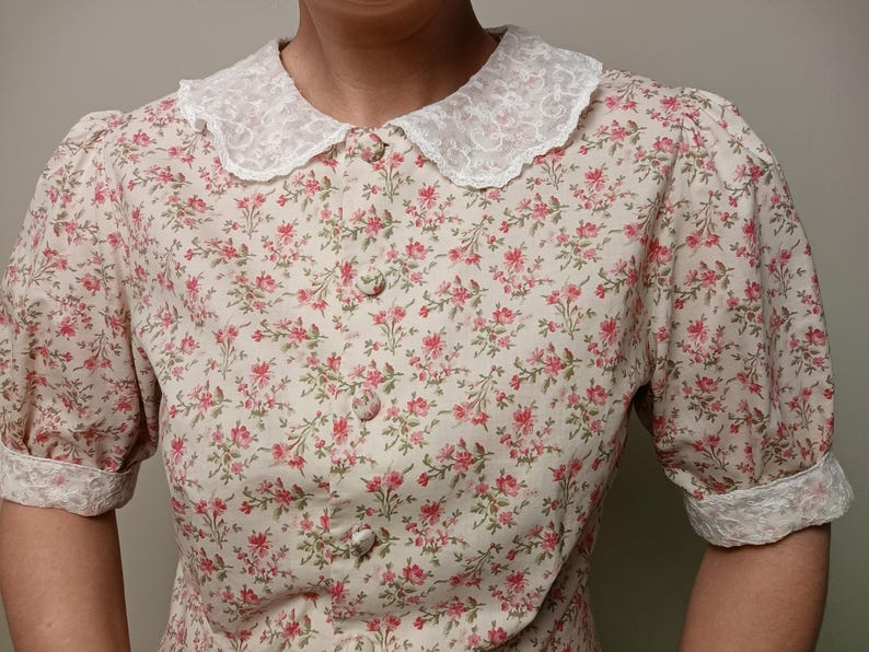 K&ouml;nnte beinhalten: Eine Bluse mit floralem Muster und einem wei&szlig;en Spitzenkragen und -b&uuml;ndchen. Die Bluse hat ein Muster aus kleinen rosa Blumen auf cremefarbenem Hintergrund. Sie hat kurze, gepuffte &Auml;rmel und eine Knopfleiste.