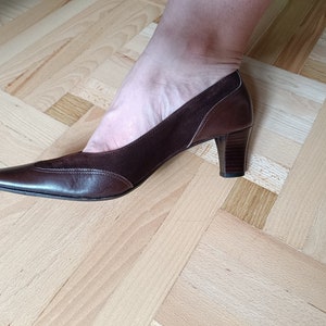 May include: A pair of brown leather heeled shoes with a pointed toe. The shoes have a suede panel on the top and a small decorative stitch detail.