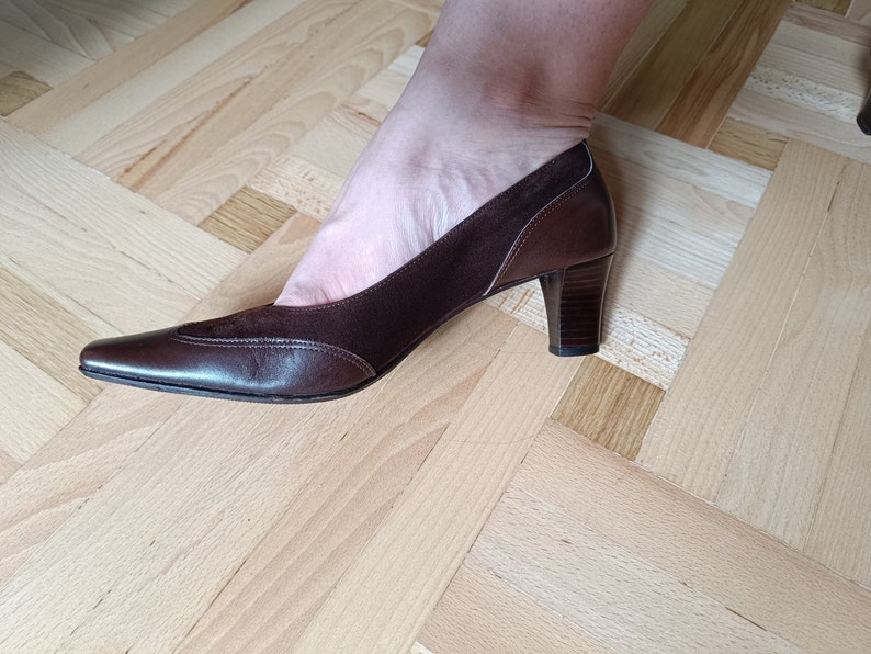 May include: A pair of brown leather heeled shoes with a pointed toe. The shoes have a suede panel on the top and a small decorative stitch detail.